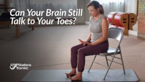 Concise version (recommended for web): "Woman seated on chair practicing toe mobility exercise in bright Pilates studio, focusing downward at her bare feet on blue mat