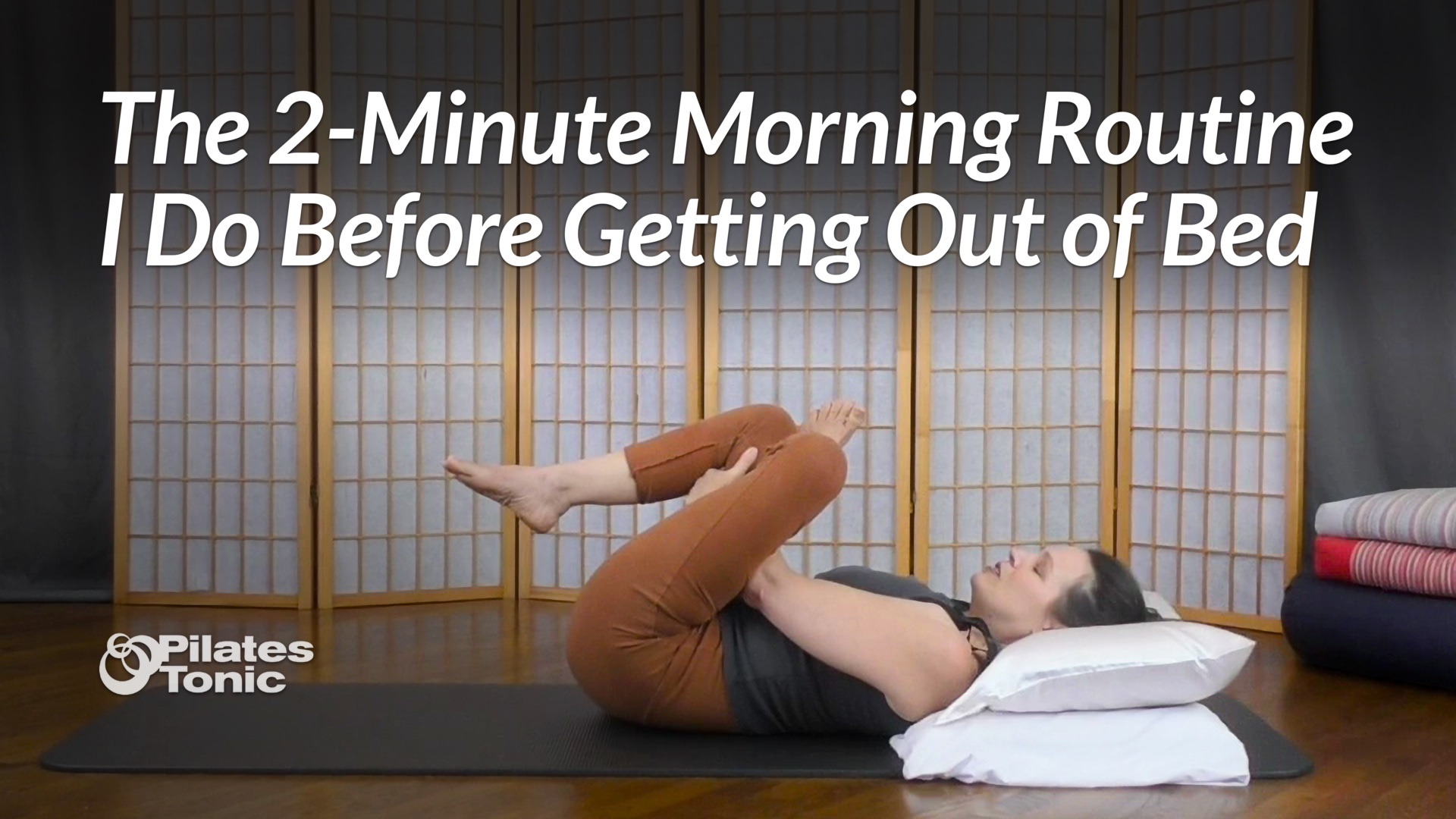 Sydney lying on a mat with her head on pillows, hugging one knee to her chest while extending the other leg in a gentle morning stretch.